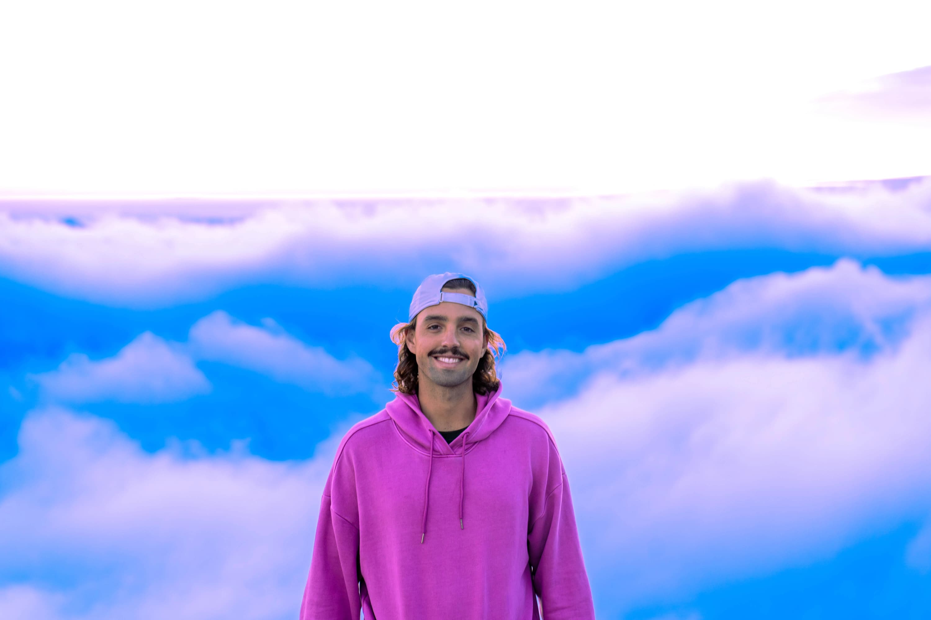 Cam smiling above a sea of clouds, wearing a pink hoodie and backwards cap