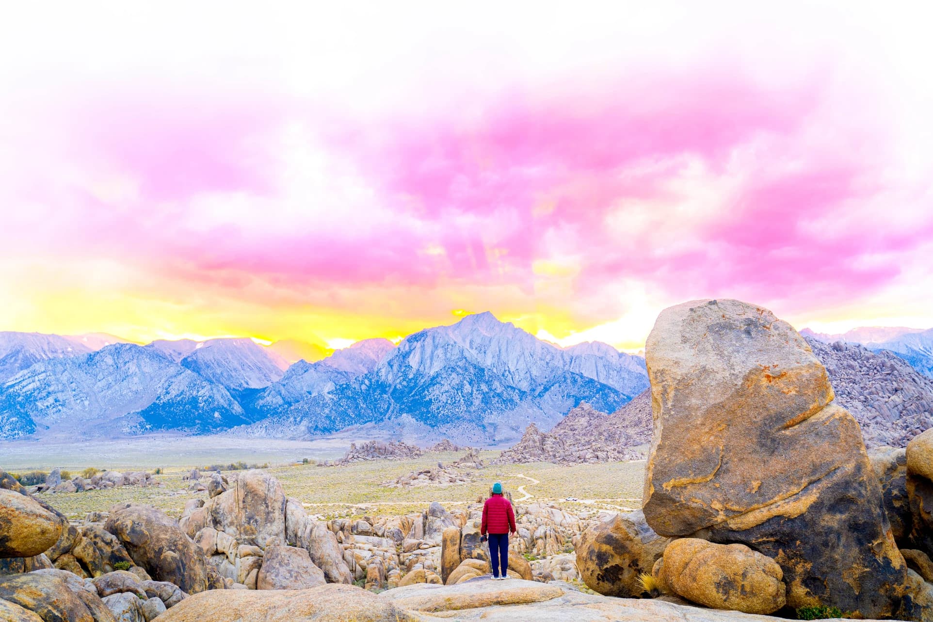 Cam standing among giant boulders facing a jagged peak under a pink sky